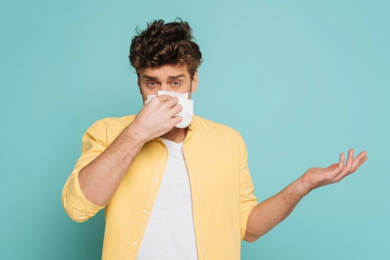 Front view of man blowing out nose with napkin, pointing with hand and looking at camera isolated on blue